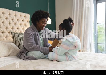 Mamma e figlia afroamericane felici sedute sul letto spazzolando i capelli a casa Foto Stock