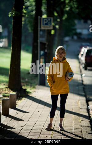 Una donna con un tappetino yoga cammina in un parco cittadino. Foto Stock