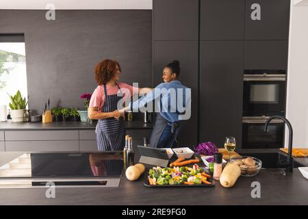 Coppia felice e variegata in grembiuli che prepara il pasto divertendosi a ballare in cucina Foto Stock