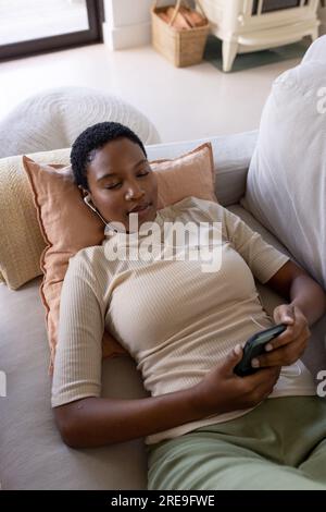 Donna afro-americana rilassata che indossa auricolari sdraiati sul divano, utilizzando lo smartphone Foto Stock