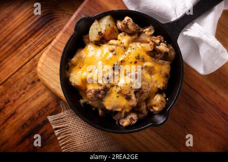 Ricetta fatta in casa per patate con bistecca e formaggio fuso, condita e servita in una padella di ghisa, su un tavolo rustico di legno. Vista superiore tabella. Foto Stock