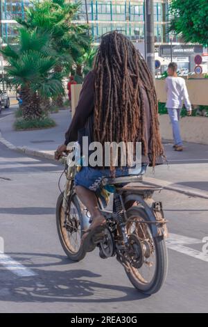 Una foto da dietro di un uomo con i capelli africani che guida una vecchia motocicletta in mezzo alla strada in una città Foto Stock