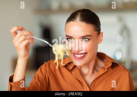 Giovane donna caucasica affamata che mangia gustosa pasta fatta in casa, tenendo in mano e guardando gli spaghetti spiedati su una forchetta Foto Stock