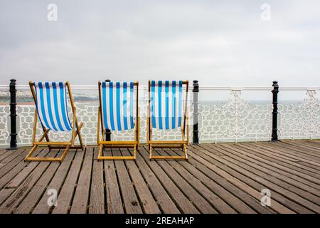 Tre sdraio a strisce blu e bianche che si affacciano sul mare sul molo di Brighton, con un cielo grigio sovrastato. Foto Stock