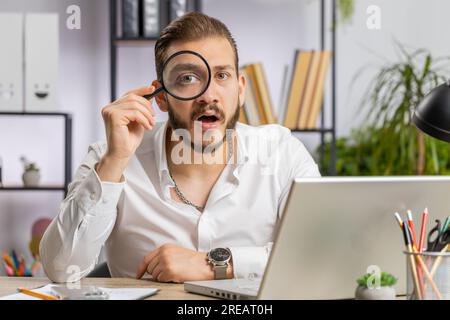 Ricercatore ricercatore scienziato uomo d'affari che lavora a casa ufficio tenendo lente d'ingrandimento vicino al viso guardando in macchina fotografica con grande zoomato divertente occhio, la ricerca, l'analisi. Uomo freelance professionista Foto Stock