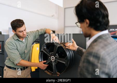 Vista posteriore del venditore professionista in tuta aziendale che parla delle caratteristiche dei cerchi in lega al cliente maschio Foto Stock