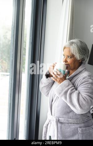 Donna birazziale anziana premurosa che indossa accappatoio e beve caffè alla finestra a casa Foto Stock