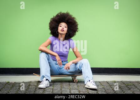 Corpo completo di giovane donna africana in acconciatura afro e abiti casual con trucco seduto sullo skateboard su un pavimento terrazzato e guardando la macchina fotografica aga Foto Stock