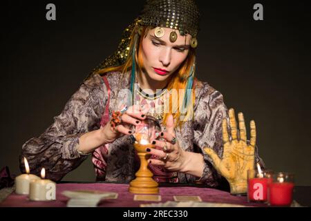 Giovane indovina femminile concentrata con lunghi capelli di zenzero in abito e bandana seduta al tavolo con candele brucianti carte e palla di cristallo Foto Stock