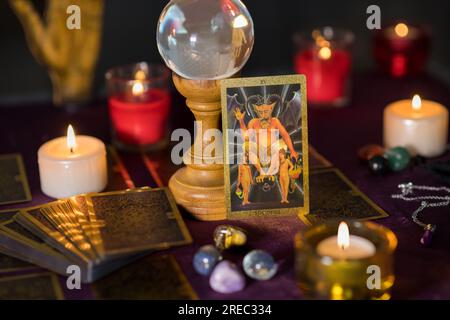 Set di candele incendiate con tarocchi del diavolo e pallina di vetro trasparente con pietre colorate lisce su un piedistallo di legno posizionato sul tavolo del indovino Foto Stock