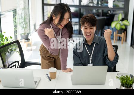 Due giovani e felici impiegati asiatici guardano lo schermo di un notebook, rallegrandosi e celebrando il successo del loro progetto in ufficio Foto Stock
