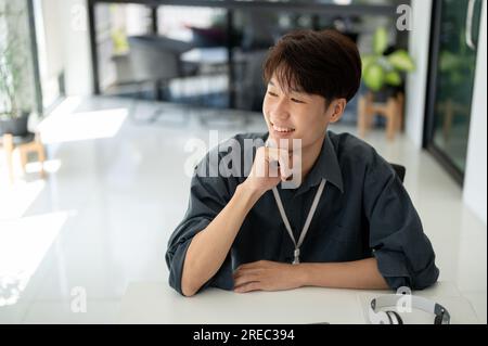 Un giovane uomo d'affari asiatico intelligente e felice guarda fuori dalla finestra, pensa e pianifica qualcosa o sogna ad occhi aperti sulla sua carriera di successo Foto Stock