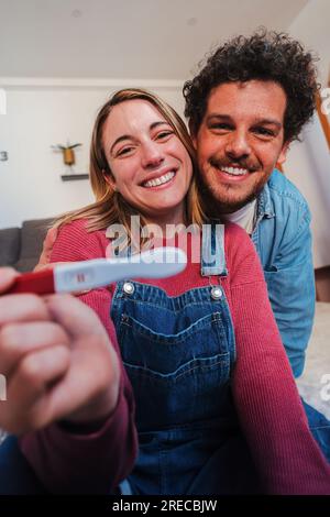 Ritratto verticale di una giovane coppia felice che sorride entusiasta di mostrare alla fotocamera il test di gravidanza positivo. Marito e moglie festeggiano che stanno aspettando un bambino che tiene in mano un predittore. Foto di alta qualità Foto Stock