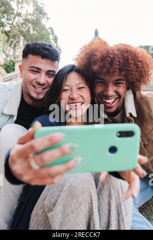 Ritratto verticale di un gruppo di amici adolescenti multirazziali che si divertono a scattare un selfie con un telefono cellulare. Giovani felici che sorridono e guardano video divertenti o fanno una videochiamata con un cellulare. Foto di alta qualità Foto Stock