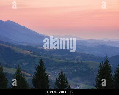 tramonto sulle montagne dei carpazi. lontana valle rurale con dolci colline tra pendii boscosi in un'atmosfera nebbiosa. cielo rosa Foto Stock