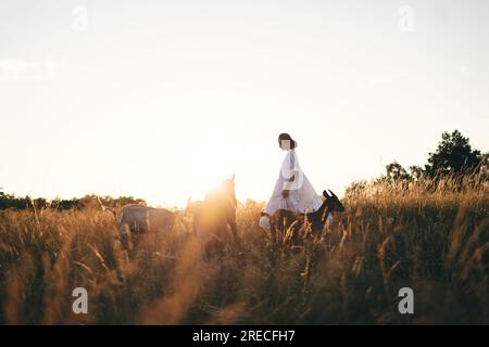 Una giovane donna in abito bianco cammina con le capre sul prato al tramonto. Attraente contadina che dà da mangiare alle sue capre con la sua piccola impresa biologica Foto Stock