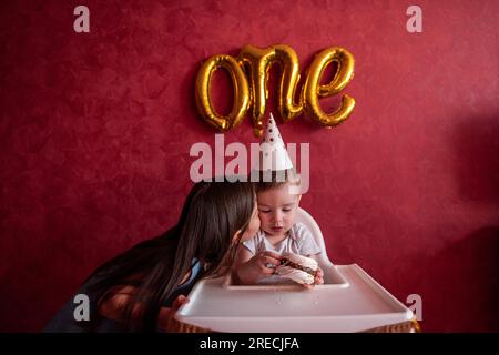 La sorella maggiore si congratula con il fratello minore per il suo primo compleanno. La ragazza bacia gli abbracci da bambino. I bambini si divertono su sfondo rosso con la lamina B. Foto Stock