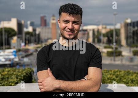 Giovane ragazzo bello sorridente si rilassò e guardò la macchina fotografica per strada con le braccia incrociate. Uomo sicuro e felice rilassato e che fissa la fotocamera in piedi Foto Stock