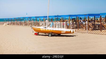 Piccola barca - barca a vela, marrone chiaro di colore arancio scuro. Si trova su una spiaggia sabbiosa, con ombrelloni blu sullo sfondo. Formato: panorama Foto Stock
