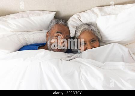 Ritratto di una felice coppia birazziale anziana sdraiata nel letto coperto da biancheria da letto a casa Foto Stock
