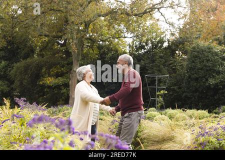 Felice coppia birazziale anziana che balla con fiori intorno a loro in giardino a casa Foto Stock