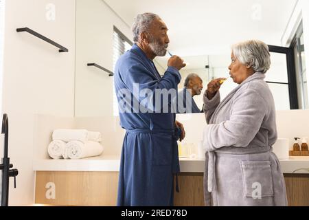Coppia birazziale anziana che indossa gli accappatoi e si lava i denti in bagno a casa Foto Stock