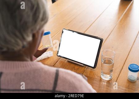 Donna anziana birazziale che ha videochiamata con medico su tablet con spazio per la copia, tenendo pillole a casa Foto Stock