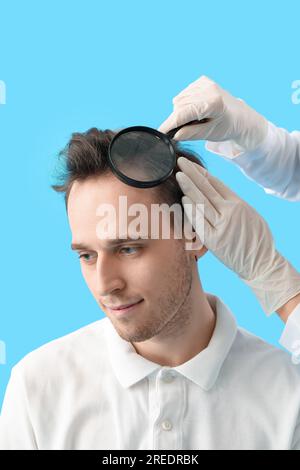 Medico con lente d'ingrandimento che esamina i capelli del giovane uomo su fondo blu, primo piano Foto Stock