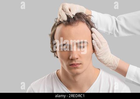 Medico che esamina giovane uomo con problemi di perdita di capelli su sfondo grigio, primo piano Foto Stock