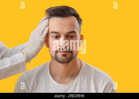 Medico che esamina i capelli del giovane su sfondo giallo, primo piano Foto Stock