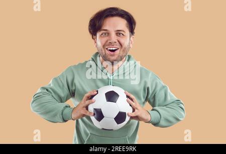 Tifoso di calcio felice giovane eccitato che tiene la palla in mano su sfondo beige. Foto Stock