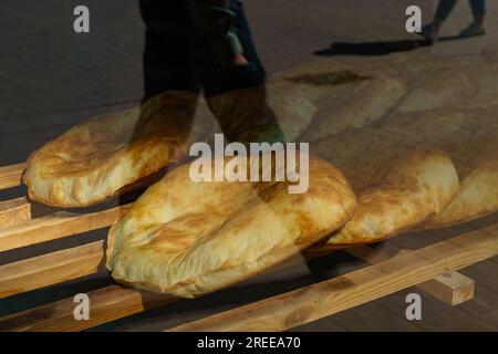 Il pane pita appena sfornato si trova su una griglia di legno in una panetteria. Vista dalla strada attraverso una vetrina di vetro che riflette le persone che passano. Foto Stock