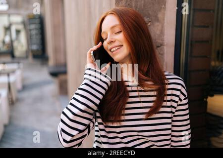 Ritratto in primo piano di una giovane donna rilassata che parla al telefono cellulare seduto sopra il davanzale con gli occhi chiusi. Felice donna rossa sorridente che cammina Foto Stock