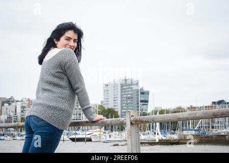 giovane donna latina turistica avventurosa in maglione grigio e pantaloni jean blu, si trova in piedi appoggiandosi alla ringhiera del molo sul fiume, visitando Foto Stock
