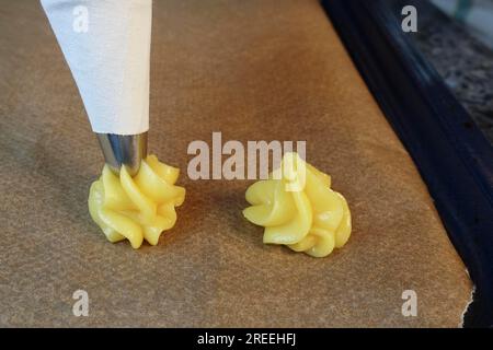 Cucina sveva, preparazione di piccoli batuffoli di panna, pressare la pasta choux cruda attraverso il sacchetto di piping su carta da forno, eclairs, profiteroles, pasticceria in piping Foto Stock