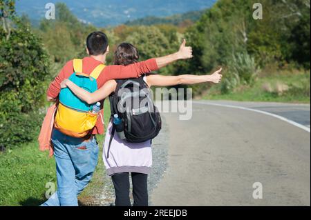 Giovani coppie, escursionisti con zaino in spalla, autostop su una strada collinare in una giornata di sole. Concetto di zaino autostop Adventure. Vista posteriore Foto Stock