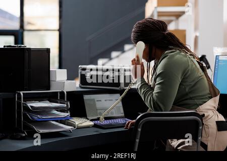 Responsabile della preparazione dell'ordine che discute della divisione di spedizione con il cliente remoto presso il telefono fisso in magazzino. Dipendente afroamericano che analizza la lista di controllo delle merci su un computer portatile nel magazzino Foto Stock