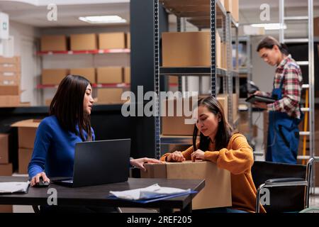 Responsabile della consegna del magazzino che verifica la qualità dell'imballaggio dei pacchi mentre lavora con il commissionatore su sedia a rotelle. Gli operatori addetti alla spedizione che preparano scatole di cartone con merci per la distribuzione Foto Stock