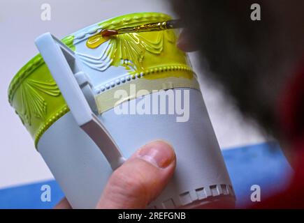 25 luglio 2023, Berlino: Una tazza della serie Kurland è parzialmente ricoperta di lacca al Königliche Porzellan-Manufaktur di Berlino per creare uno sfondo non smaltato. La porcellana è stata realizzata a mano al KPM Berlin per 260 anni. Più di 200 dipendenti lavorano attualmente al progetto "Handmade in Berlin" presso la manifattura fondata da Federico il grande. Occorrono 29 fasi di lavoro, eseguite in 14 giorni lavorativi da 25 operai della manifattura, per produrre una tazza di porcellana bianca con il reclamo KPM Berlin. Con 10 controlli di qualità, ogni oggetto viene ordinato a mano. Foto: Jens Kalaene/dpa Foto Stock