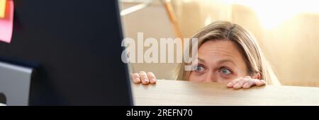Donna si nasconde dietro la scrivania dell'ufficio e guarda il monitor del computer. Foto Stock