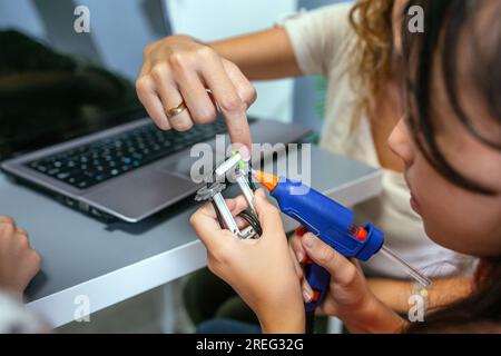 Insegnante che aiuta gli studenti a utilizzare la pistola per colla a caldo su pezzi di macchine in classe di robotica Foto Stock