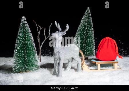 Carta di Natale creativa. Un capriolo d'argento brillante giocattolo che trasporta una slitta con una borsa di regali attraverso la foresta innevata tra gli alberi di Natale di notte. Foto Stock