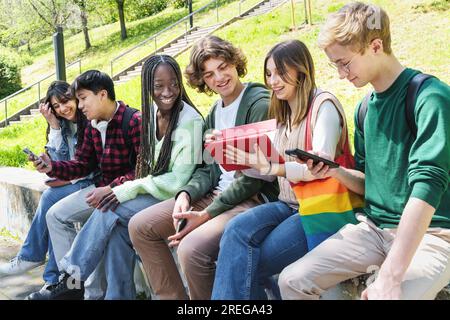 Un gruppo di adolescenti multiculturali seduti su un muro nel parco scolastico, impegnati in conversazioni e utilizzando smartphone e tablet. Foto Stock