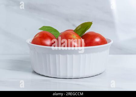 Pomodori rossi e basilico verde per un'insalata caprese su un piatto/recipiente bianco su un tavolo e uno sfondo in marmo Foto Stock