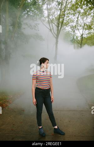 Giovane donna in piedi su un sentiero nebbioso in un parco che guarda lontano Foto Stock