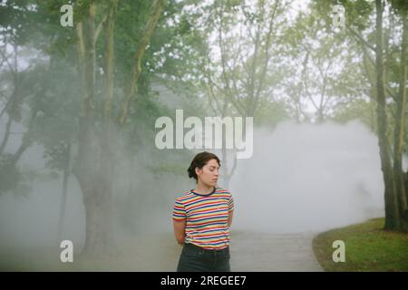 Giovane donna con occhi abbassati in piedi su un sentiero in un nebbioso parco Foto Stock