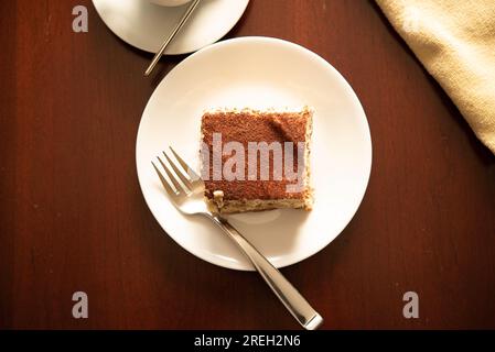 Fetta di tiramisù tradizionale italiano su piatto bianco e tavolo in legno con forchetta e tazza di caffè. Foto Stock