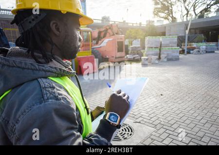 il supervisore tecnico maschio nero, indossando un casco di sicurezza e un giubbotto riflettente, sta contando e prendendo nota dei materiali del cantiere, indu Foto Stock