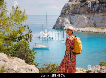La donna asiatica in cappello si affaccia sulla baia azzurra del mar Mediterraneo. Concetto di viaggio e vacanza. Baia di Anthony Quinn con acqua cristallina a Rodi Foto Stock