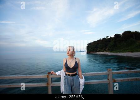 Graziosa donna bionda con camicia bianca e canotta in piedi sul molo sullo sfondo del mare, del cielo nuvoloso e della costa con alberi verdi Foto Stock